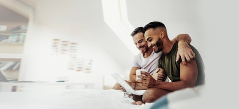 one man hugs another man as they have their laptop open smiling drinking a cup of coffee
