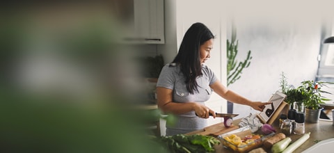 Woman scrolls on her tablet while trying to cut onions surrounded by vegetables on the counter