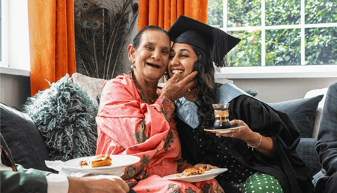 Person in graduation attire sitting with an older adult on a couch during a gathering at home.